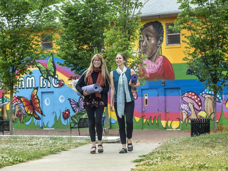 two women walking along a sidewalk with their yoga mats in front of a building with a large colorful mural of a little girl blowing bubbles, rainbows, mushrooms, butterflies, and lady bugs painted on its side
