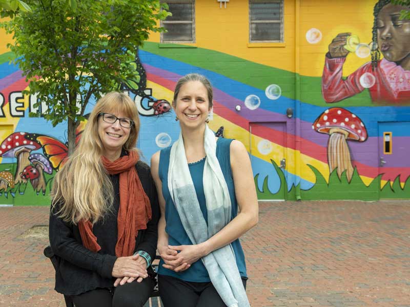 two women standing together smiling in front of a building with a large colorful mural of a little girl blowing bubbles, rainbows, mushrooms, and lady bugs painted on its side