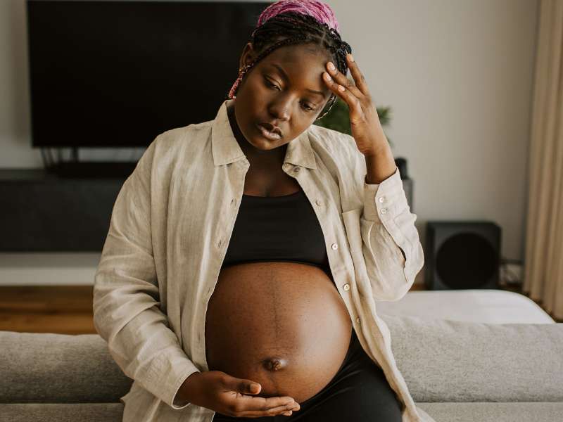 a pregnant woman holds her swollen belly with one hand and has the other hand on her forehead with her eyes closed as if she is struggling mentally